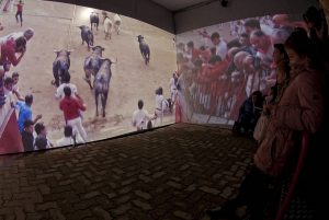 Imagen de la visita a la Plaza de Toros de Pamplona, Dos parede iluminadas con imágenes del encierro, frente a ellas un grupo de personas la observan.
